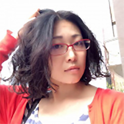 Casual portrait of a woman wearing red glasses and a red sweater, with shoulder-length wavy hair, touching her hair and looking at the camera.