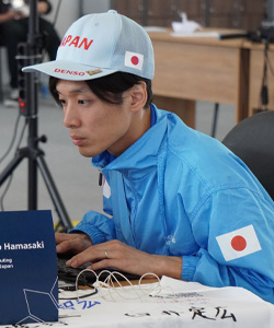 A Japanese competition participant, wearing a blue jacket and cap with Japan flag emblems, works at a computer during an event. The individual is focused on the screen, sitting at a desk with a name placard and small models.