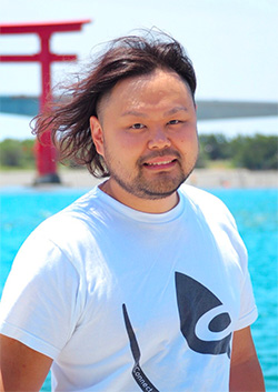 An outdoor portrait of a man with medium-length hair and a beard, standing near water with a red torii gate in the background. The image is bright and colorful, and the man is wearing a white t-shirt with a bold graphic design.