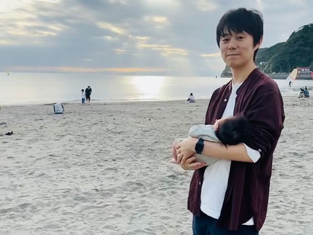 A man holding a baby while standing on a sandy beach at sunset. The sky is cloudy with light shining through, and the ocean and distant people are visible in the background.