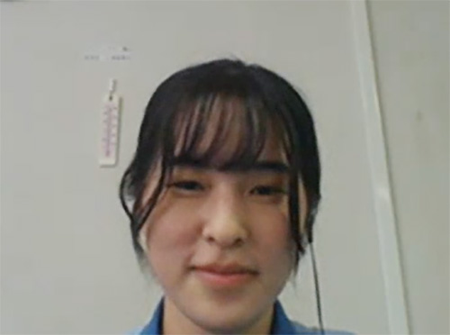 A woman with dark hair and bangs is smiling indoors, with a light background and part of a thermometer visible on the wall behind her.