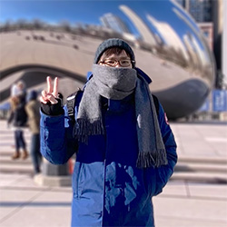 A person wearing a blue winter coat, gray scarf, and knit hat poses with a peace sign in front of Cloud Gate (The Bean) in Chicago.