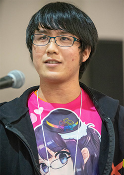 A person with glasses speaking at an event, wearing a bright anime-themed shirt, with a microphone visible in the background.