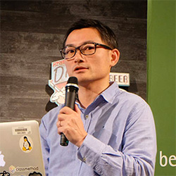 A person holding a microphone and speaking at an event, standing in front of a laptop with stickers and a green backdrop. The individual is wearing glasses and a light blue shirt. Event signage is partially visible in the background.