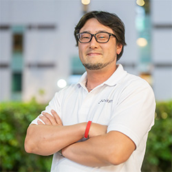 Outdoor portrait of a man in a white shirt and glasses, arms crossed, with greenery and buildings in the background, 2023.