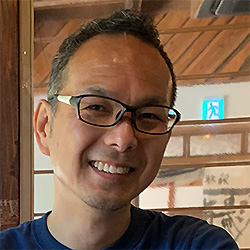 A portrait photo of a smiling person with glasses, taken indoors with wooden interior elements visible in the background.