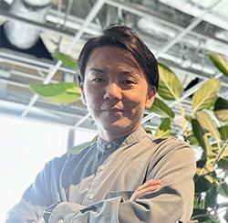 A person standing indoors with arms crossed, with plants and modern ceiling structures in the background.