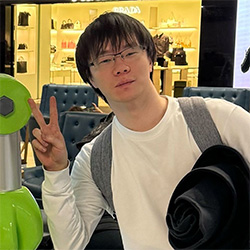 A person with short hair and glasses makes a peace sign with their fingers while carrying a hat and wearing a backpack. They are casually dressed and appear to be in a shopping area.
