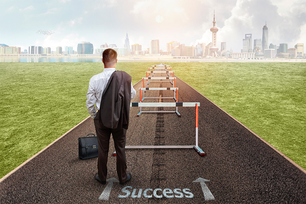 A businessman stands at the start of a track with hurdles labeled 'Success', with a cityscape visible in the background, symbolizing overcoming obstacles on the journey to achievement.