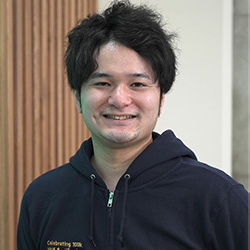 A casual portrait of a smiling person with short dark hair wearing a dark hoodie, photographed in 2023. The background features wooden and light-colored panels.