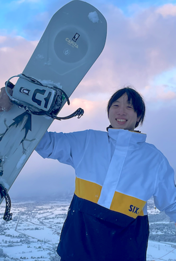 A person smiling with a snowboard on a snowy mountain, wearing a winter jacket. The scene features a winter landscape with snow and a colorful sky in the background.