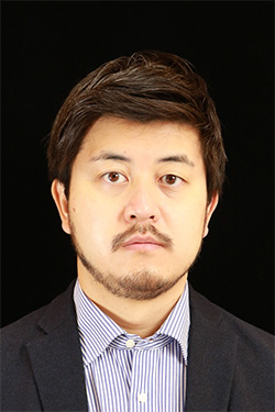 Portrait of a man in formal attire featuring a dark suit jacket and striped shirt, set against a black background.