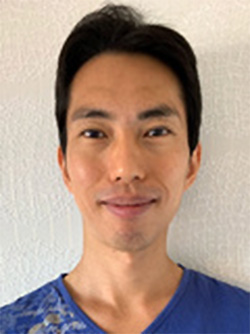 A portrait of a person smiling, wearing a blue shirt, standing against a light-colored textured wall.