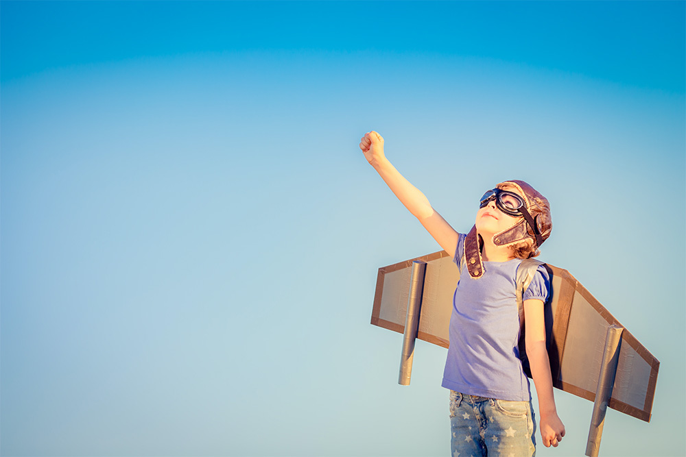 A child wearing cardboard wings and aviator goggles pretends to fly, raising one arm to the sky, against a bright blue sky background.