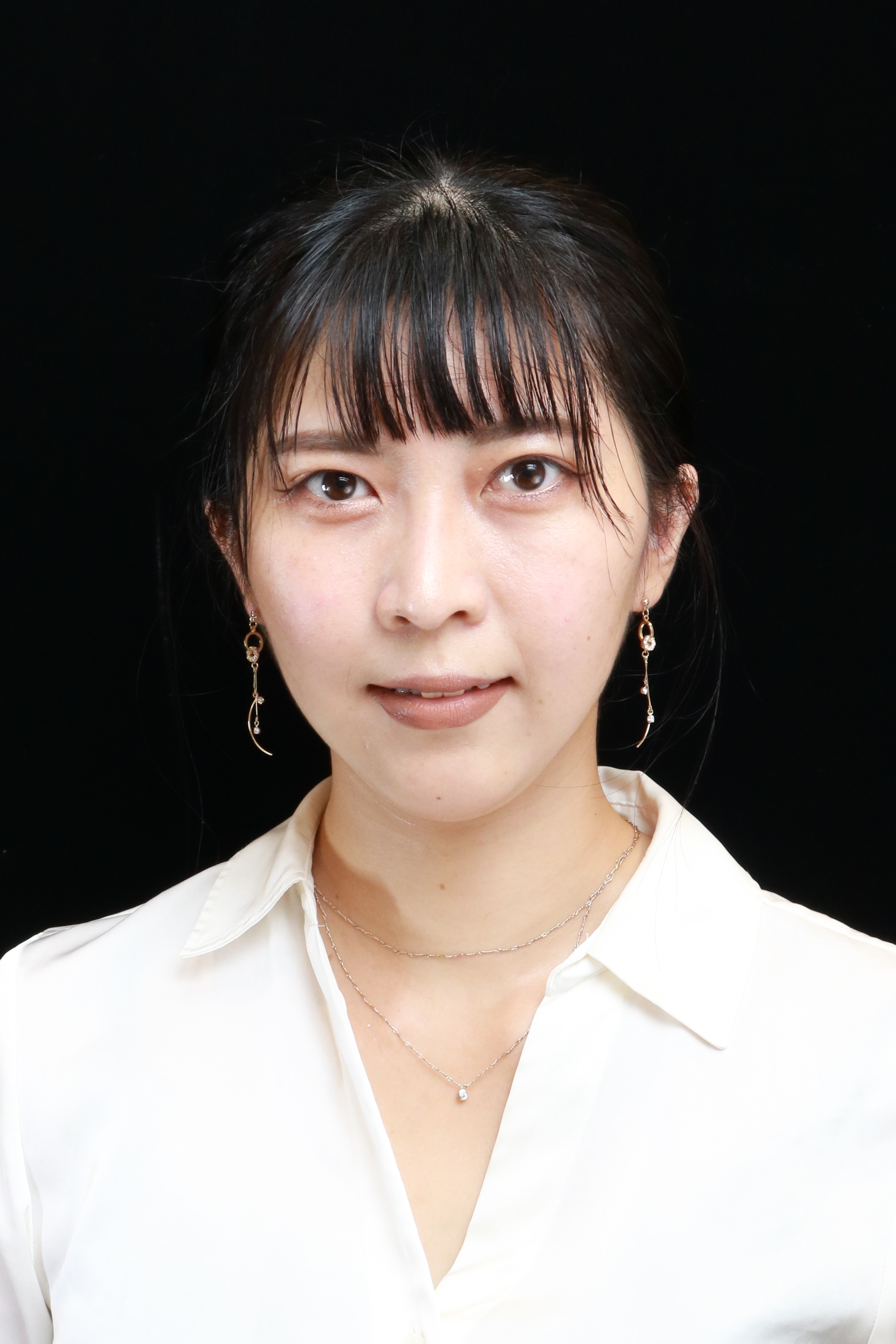 A portrait photograph of a woman with straight dark hair, wearing a white blouse, earrings, and necklaces, set against a solid black background.