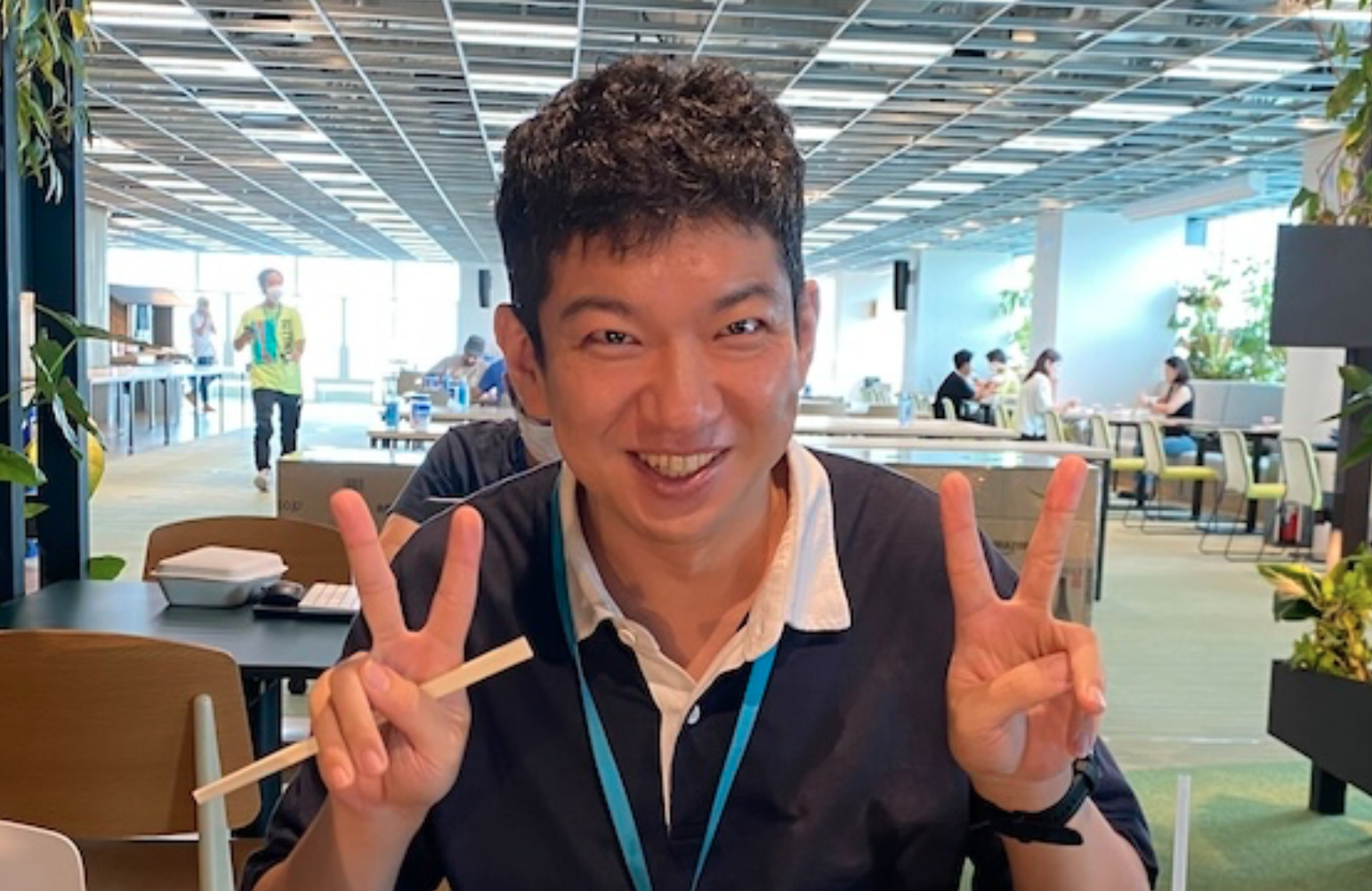 A man smiling and showing peace signs with both hands while sitting in a cafeteria. Other people can be seen in the background, suggesting a casual and friendly atmosphere.