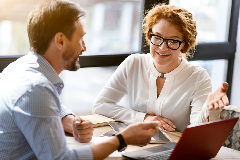 Two colleagues have a casual business discussion at a table with a laptop, notepad, and documents in a modern office setting.