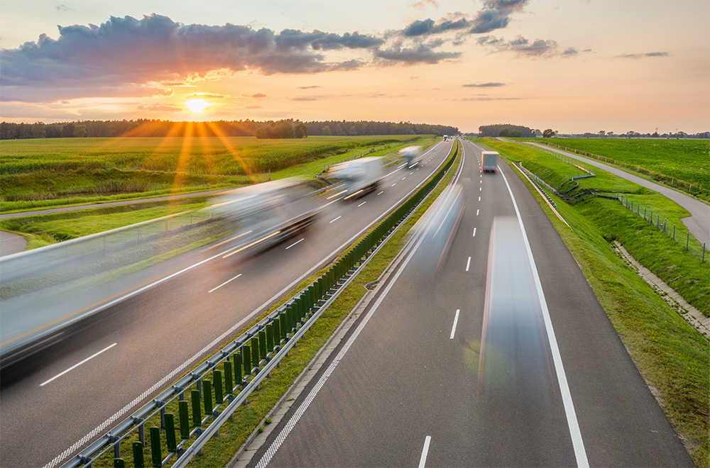 A highway with multiple lanes of traffic in motion blur as vehicles travel at high speed. The scene is set during sunset with sun rays and clouds in the sky, surrounded by green fields.