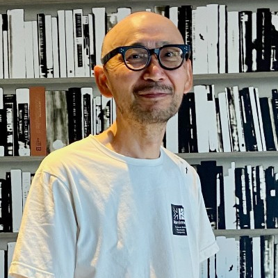 A man wearing glasses and a white t-shirt standing in front of a bookshelf background, smiling casually.