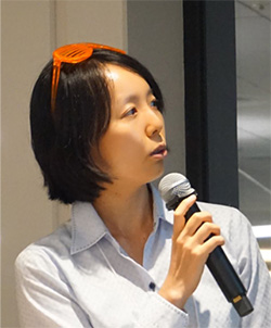 A person with short hair is speaking into a microphone, wearing a light-colored shirt and orange glasses on their head.