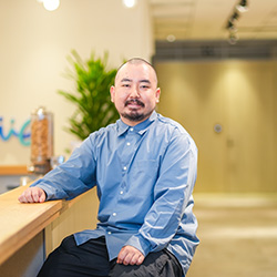 A person wearing a blue shirt sits at a wooden counter in an office setting, with blurred background and greenery.