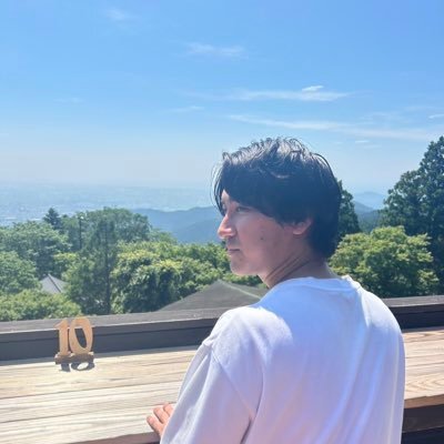 A man stands on a wooden deck looking out at scenic green mountains under a clear blue sky on a sunny day.