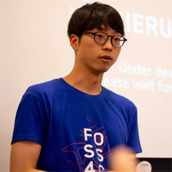 A person wearing glasses and a blue t-shirt is speaking in front of a projected screen during a presentation.