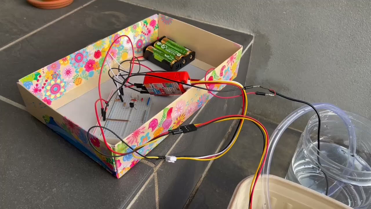 A homemade water pump circuit setup featuring electronic components on a breadboard, a battery box, and connecting wires placed inside a colorful cardboard box, with tubes leading to a water container.