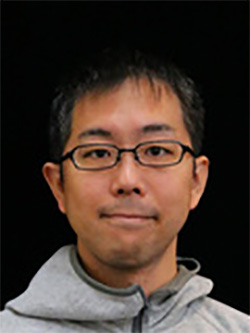 Portrait of a man with short black hair, wearing glasses and a light gray zip-up jacket, against a dark background.
