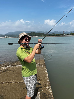 A man wearing a green shirt and hat is fishing outdoors by a river on a sunny day, with mountains and blue sky in the background.