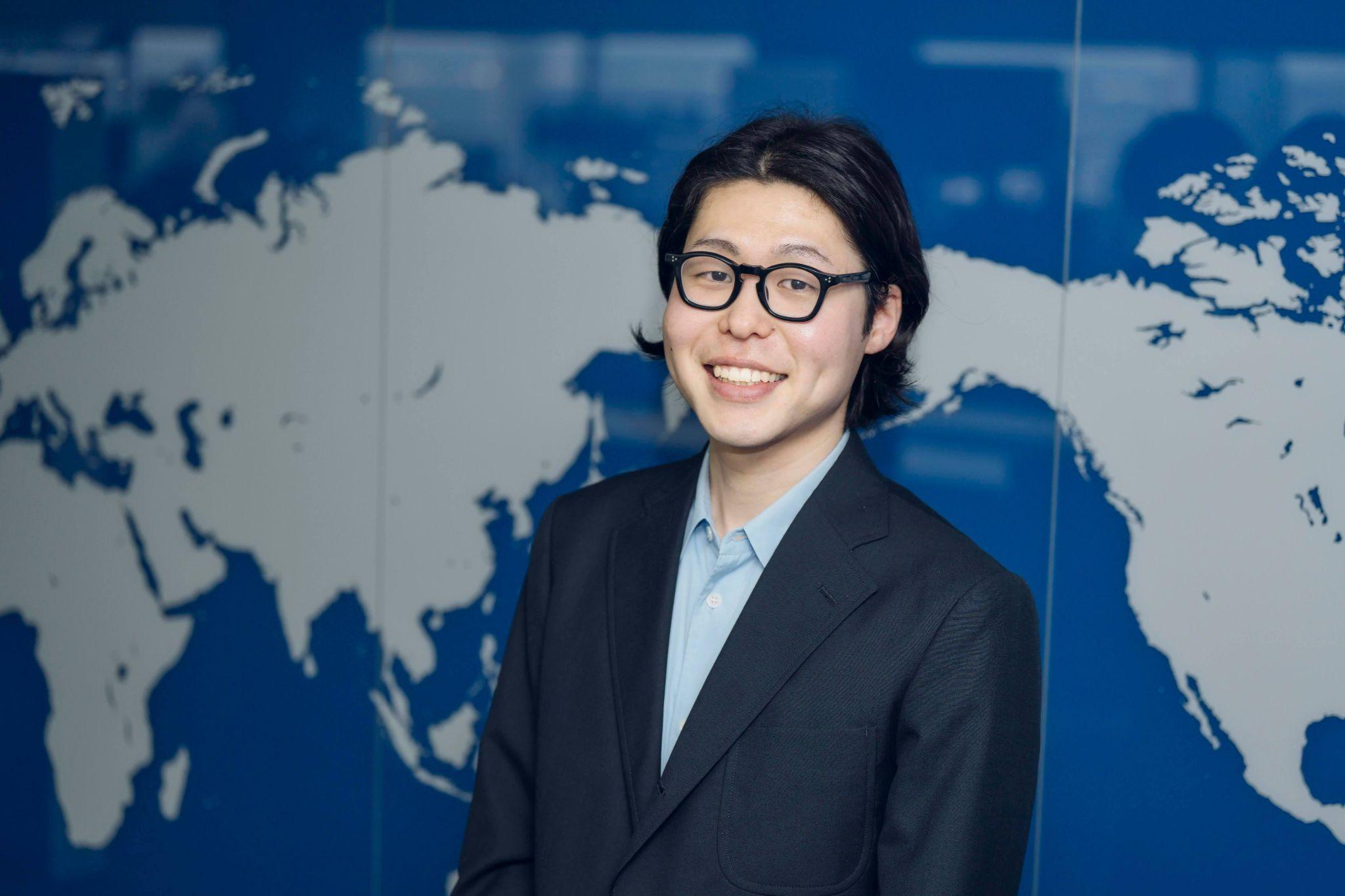 A businessman in a suit and glasses is smiling while standing in front of a world map background.