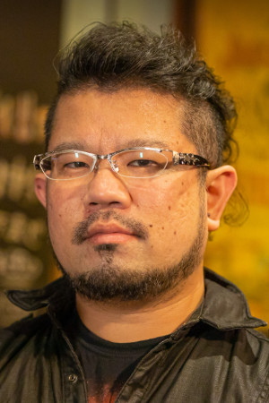 A portrait photo of an individual with short, styled hair, glasses, and facial hair, wearing a dark shirt, photographed indoors with a softly blurred background.