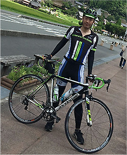 A cyclist in professional gear posing with a road bike outdoors on a paved path with greenery and buildings in the background.