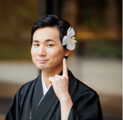 A person wearing traditional Japanese clothing with a white flower in their hair, smiling and pointing upward.