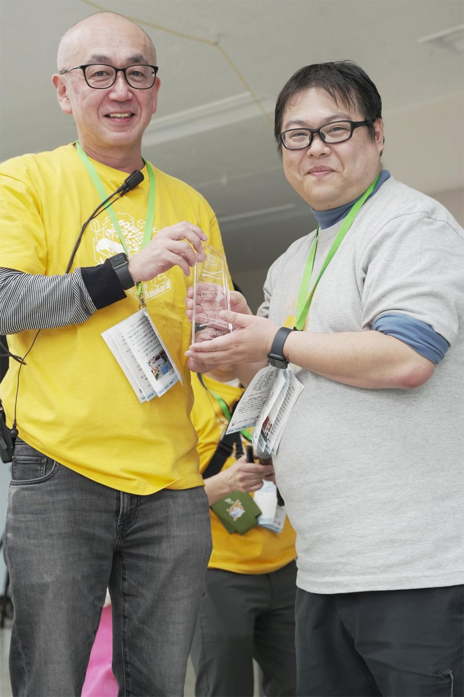 Two individuals at the AWS Samurai 2024 event present and receive an award. One person wears a yellow shirt and hands a glass award to another person in a gray shirt. Both are smiling and wearing event badges and lanyards.