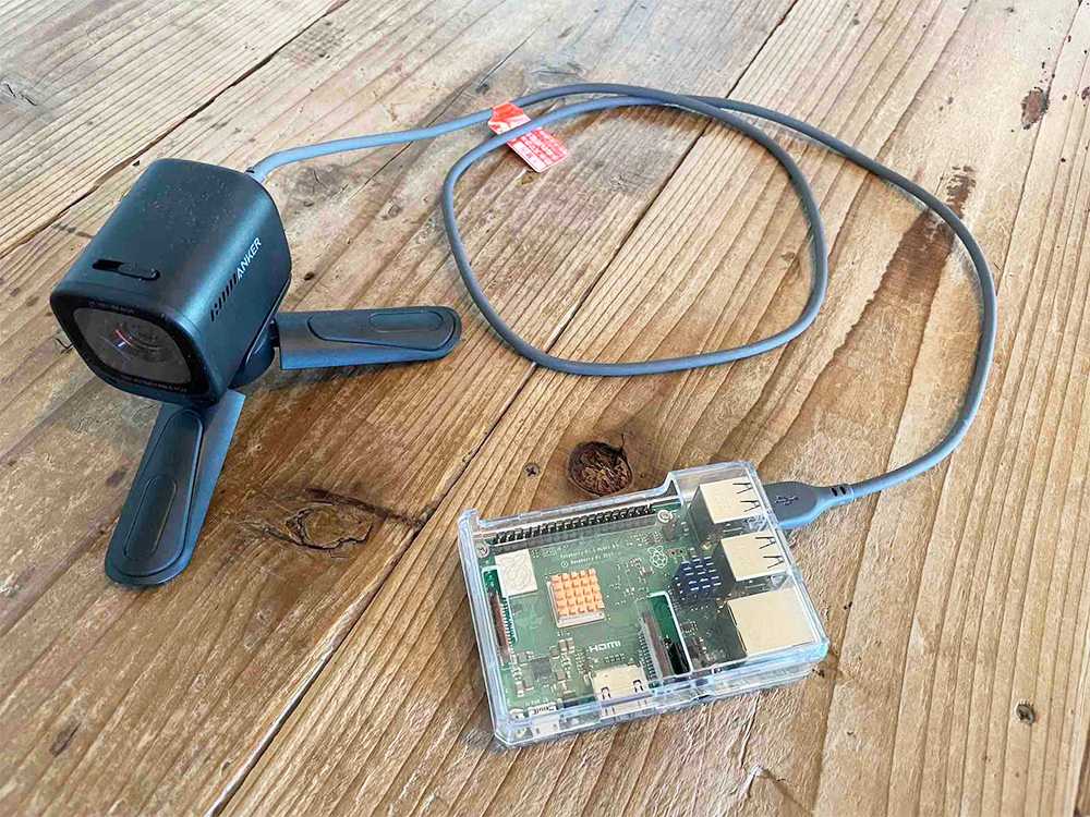 A Raspberry Pi device connected to an Anker camera with a USB cable, both resting on a wooden table.