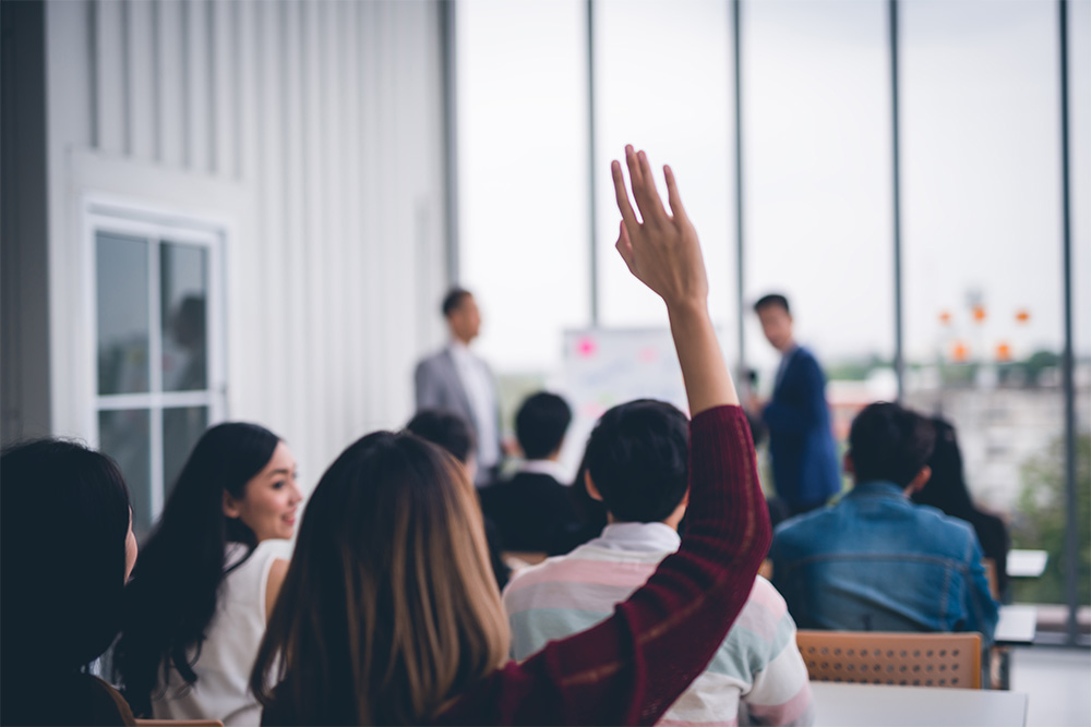 A participant raising their hand during a training session or seminar, with a group of people seated and two presenters at the front of the room.