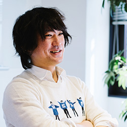 A casual office portrait of a man with medium-length dark hair wearing a white sweater with graphic prints, standing with arms crossed and smiling in a brightly lit office environment.