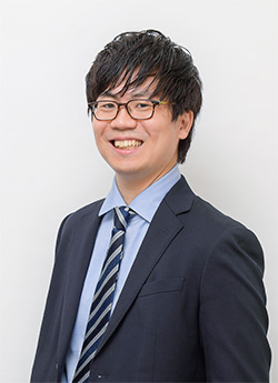 A professional business portrait of a smiling man wearing glasses and a dark suit, with a light blue shirt and striped tie, posed in front of a plain light background.
