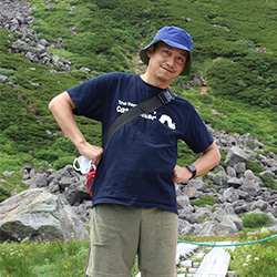 A person standing outdoors in a mountain setting, wearing a hat and casual hiking clothes, with greenery and rocks in the background.