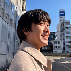 A smiling man in a light coat standing outdoors, with city buildings in the background on a sunny day.