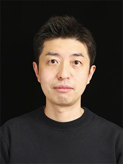 A portrait photograph of a man wearing a black shirt, standing in front of a plain black background.