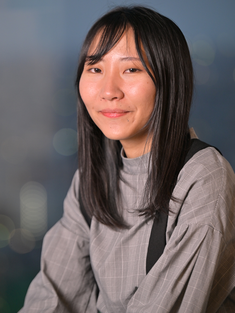 A portrait of a woman with straight dark hair wearing a gray plaid top, photographed against a softly blurred background.