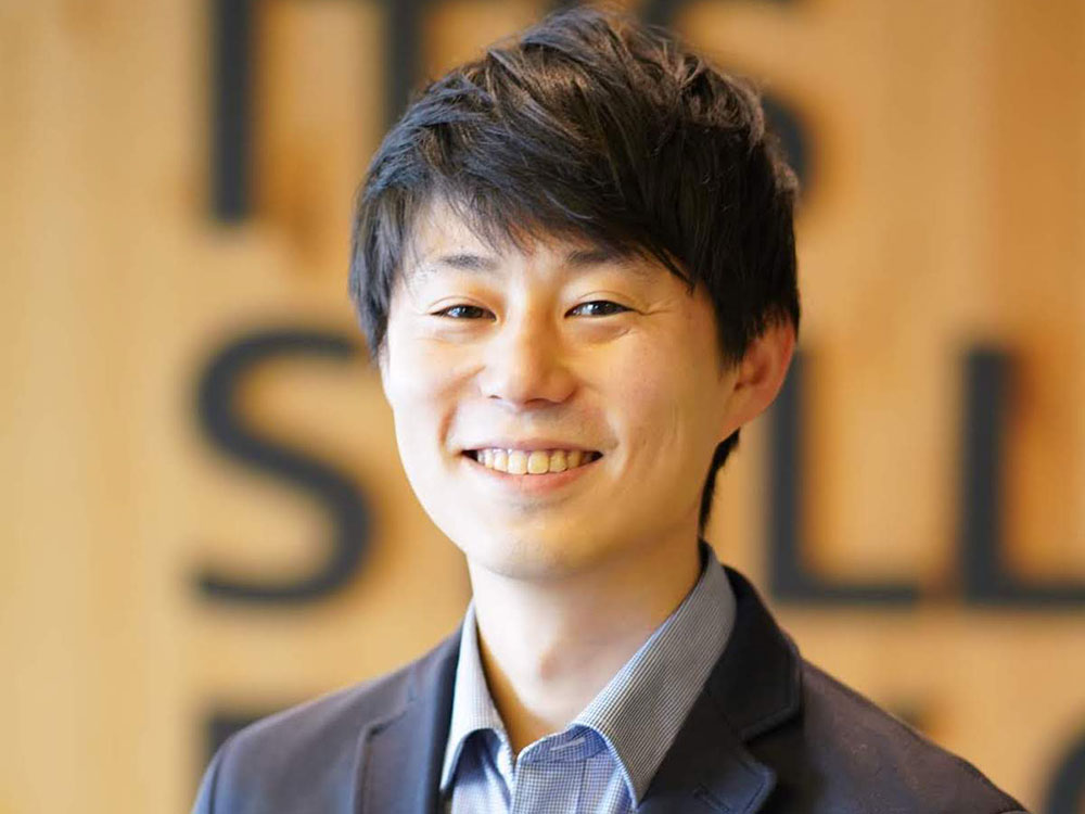 A portrait of a smiling young Asian man in business casual attire, standing in front of a blurred office background.