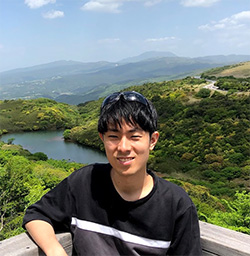 A person smiling outdoors with a scenic mountain and lake view in the background.