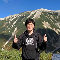 A person standing outdoors in front of a scenic mountain landscape, smiling and giving two thumbs up. The individual is wearing a dark hoodie. The weather is clear and sunny.