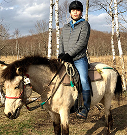 A person wearing a helmet is riding a horse in a forest clearing with bare trees in the background.