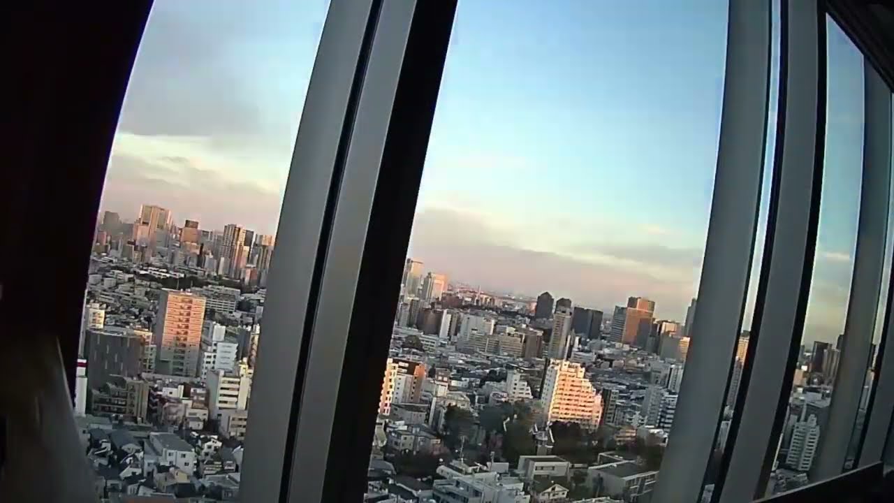 A cityscape view of modern buildings and skyscrapers, seen through large windows at sunset, with a clear sky and soft clouds in the background.
