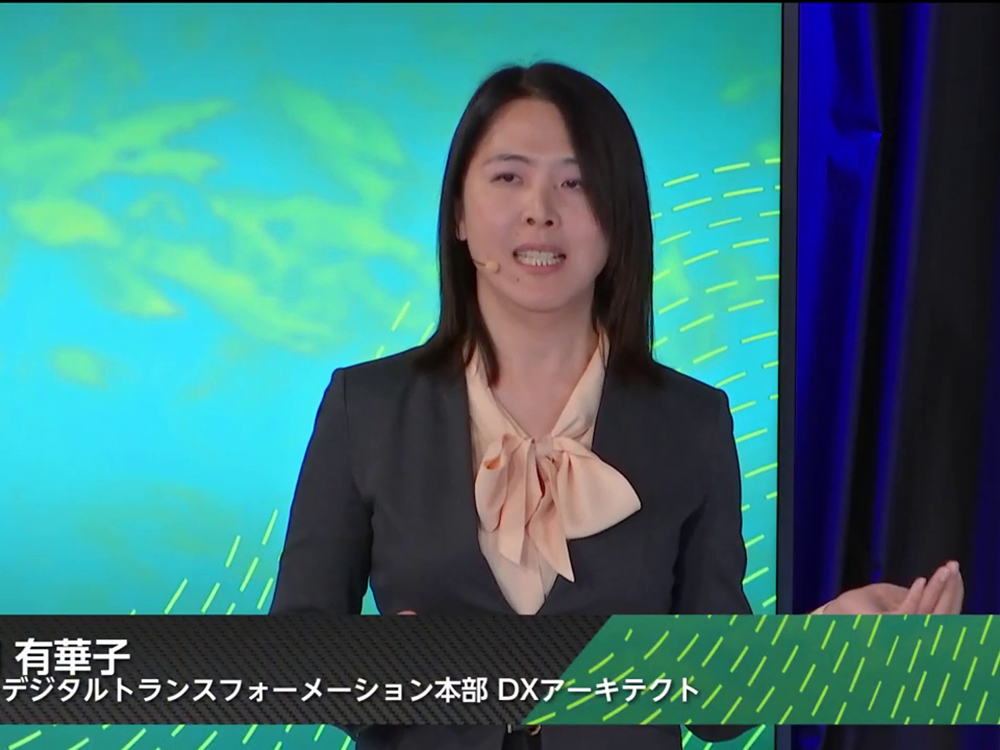 A Japanese woman in professional attire speaks on stage at a digital transformation conference, with a colorful digital background and name and title shown in Japanese text at the bottom of the image.