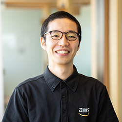 Portrait of an AWS employee wearing glasses and an AWS-branded shirt, smiling in an office setting.